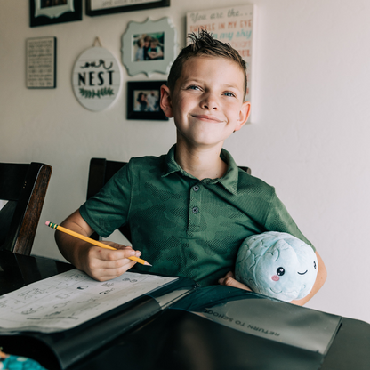 young boy wearing a green shirt sitting at a table writing in a notebook with a pencil in his right hand while holding a nerdbugs brain plushie with his left arm
