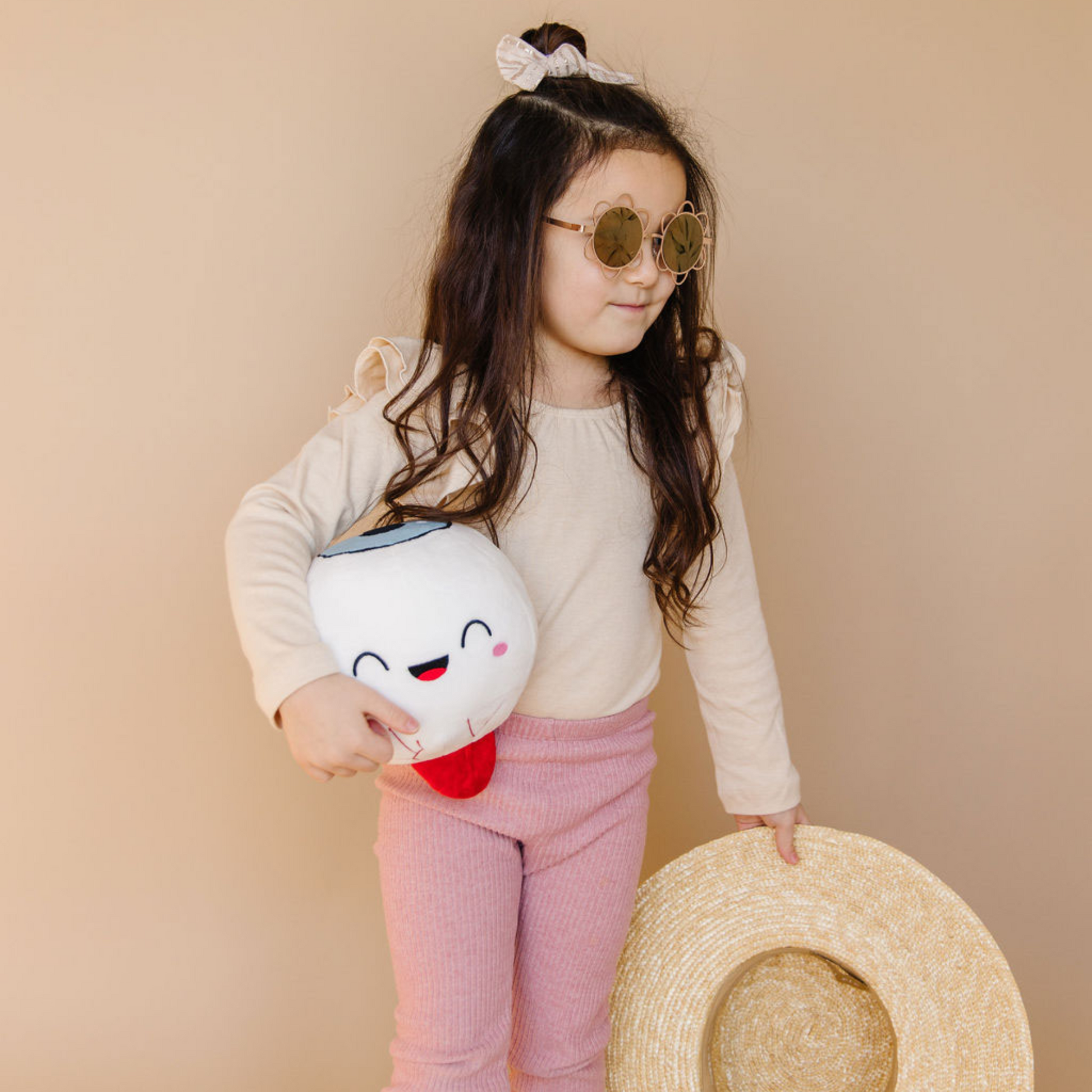 Baby girl in pink background wearing sunglass and holding a hat and an eye toy plushie. 